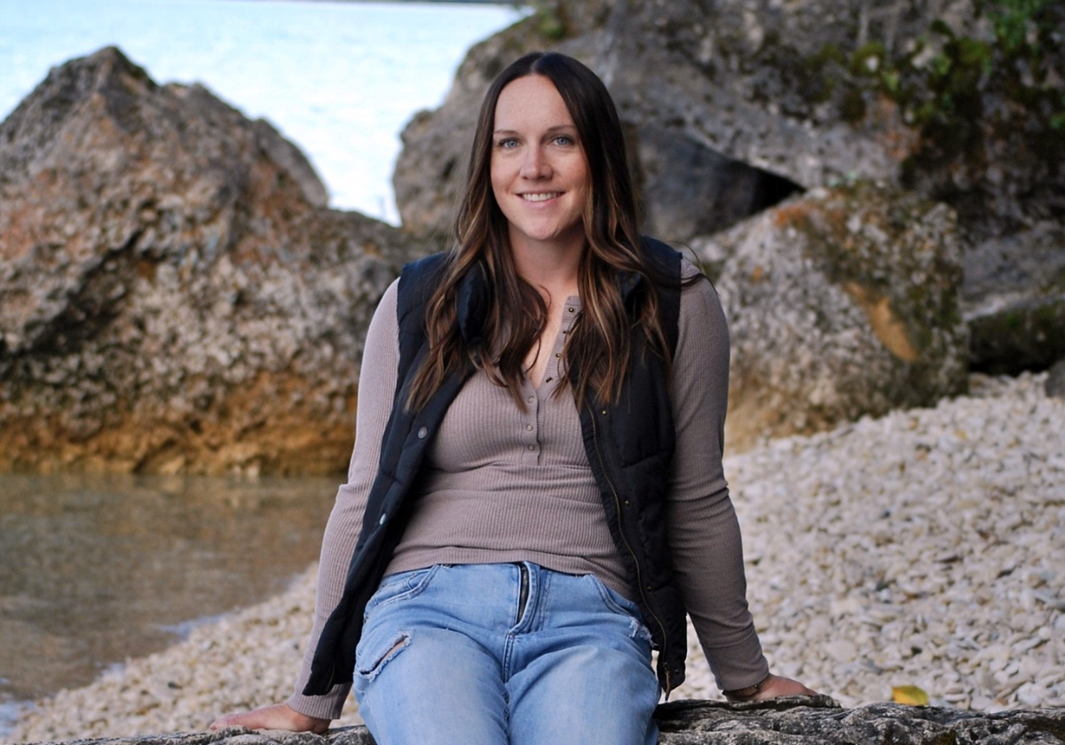 Bachelor of Science in Nursing Spring 2026 female graduate Jalene Dease sits smiling on a rock with larger rocks and a body of water in the background. 