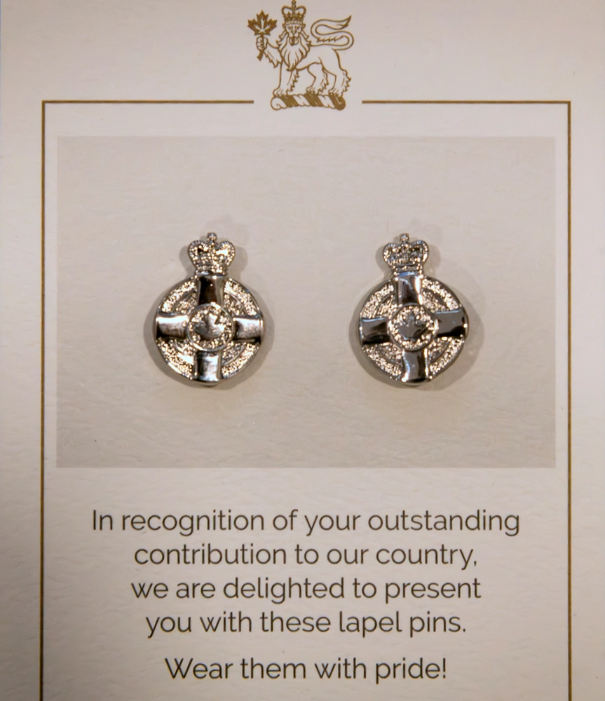 Two pins pinned on a white card with text underneath reading ‘In recognition of your outstanding contribution to our country, we are delighted to present you with these lapel pins. Wear them with pride!’