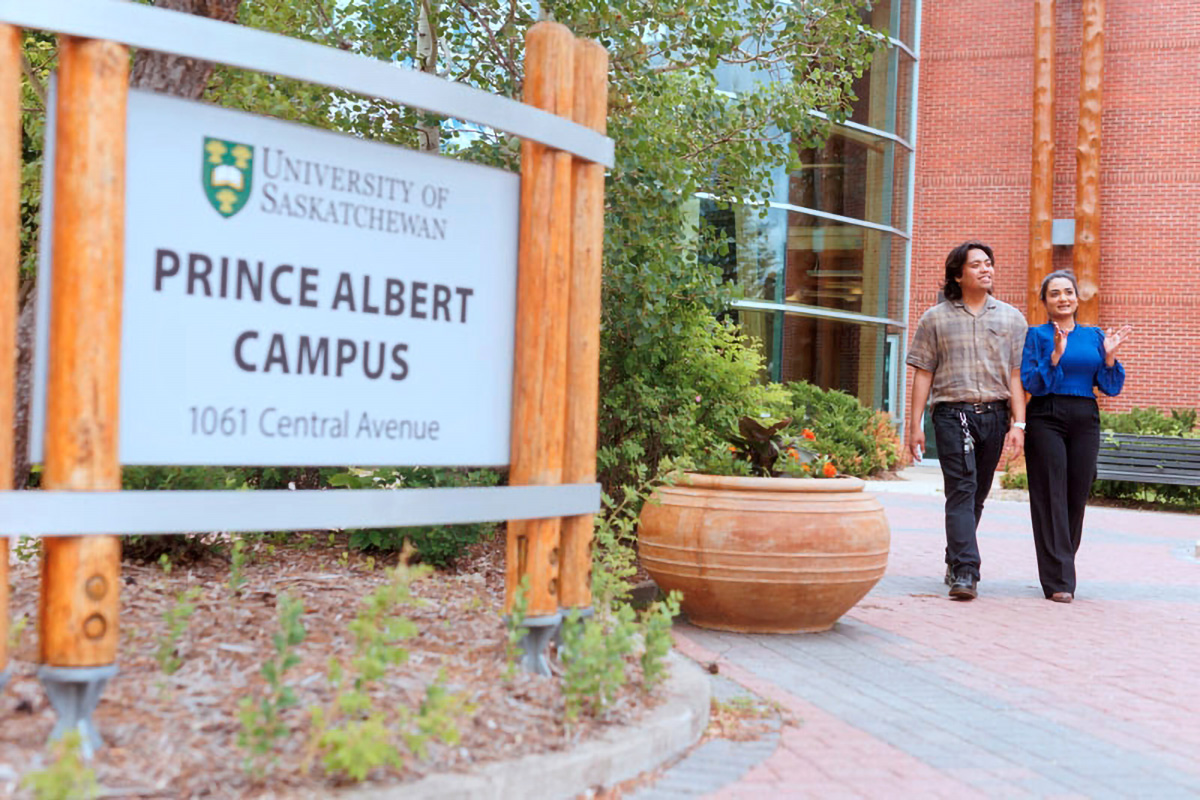 A male and a female walk outside of the glass atrium by the Prince Albert Campus sign.