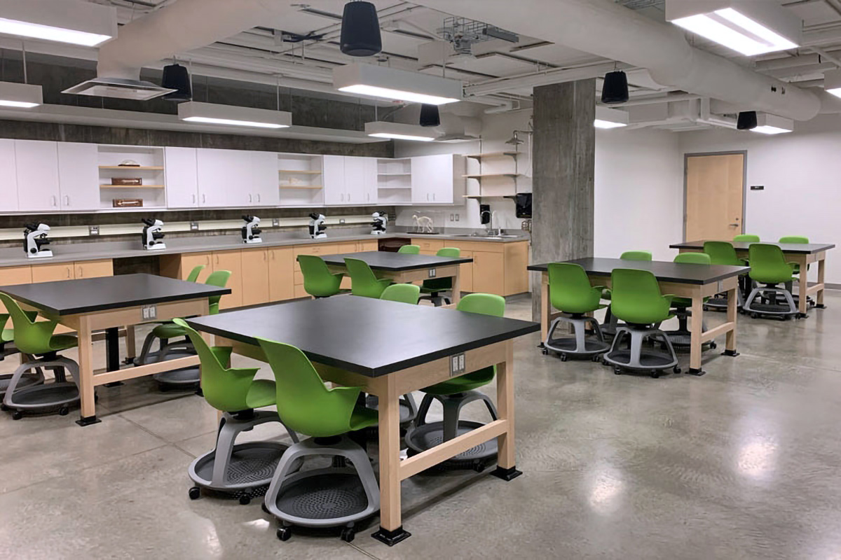 A lab setting with black tables and green chairs set up in stations of 4 and microscopes on a counter in the background.
