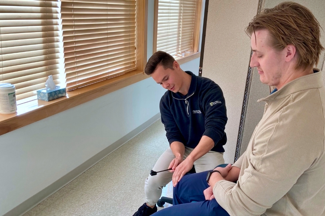 A male student assesses the reflexes of another student sitting down in a clinic like setting.