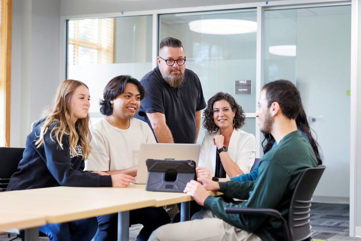 Students and staff sit at a table around a computer in a student lounge outside a student meeting space.