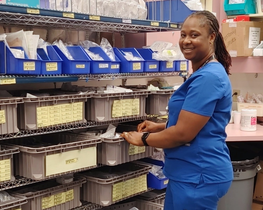 Augustina, a woman dressed in blue scrubs, stands in front of a shell with blue and tan bins of various sizes, collecting medical supplies.