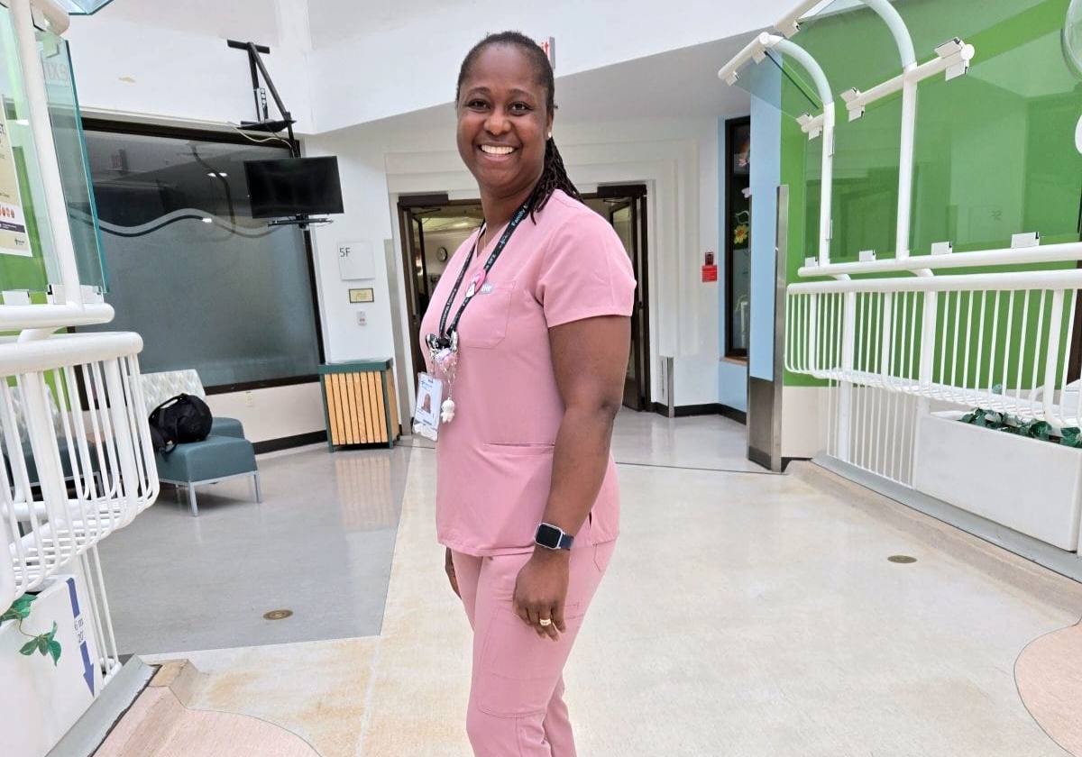 Augustina, a woman, smiling wearing light pink scrubs while standing in the middle of a hospital hallway with green walls.